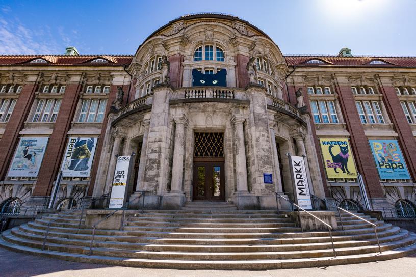 Außenansicht des MARKK bei sonnigem Wetter. Das Museumsgebäude ist im Tageslicht zu sehen, der Himmel ist klar und die Fassade wird von der Sonne beleuchtet.