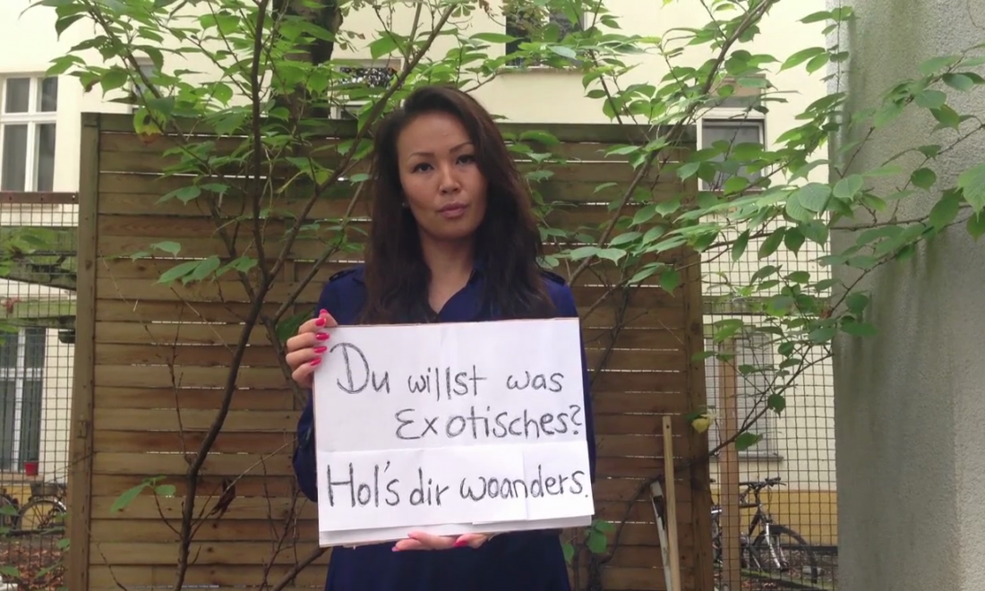 Exotik. Hols woanders – © Sun-ju Choi Eine junge Frau hält ein Schild hoch. Auf ihm steht: Du willst was Exotisches? Hol´s dir woanders